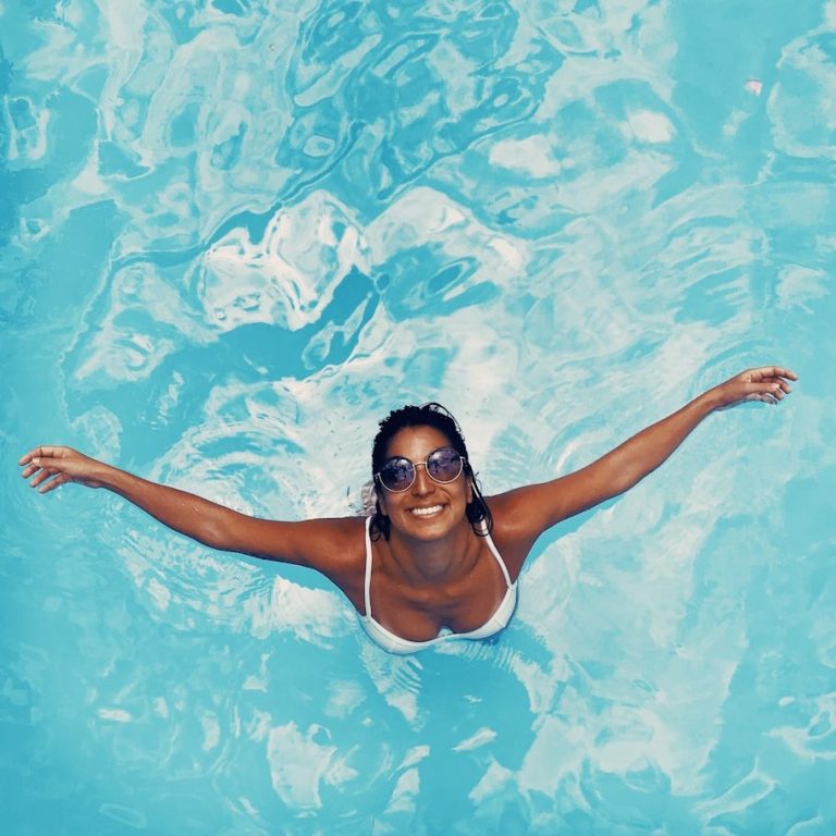 Mujer sonriendo con gafas de sol, nadando en una piscina azul clara.
