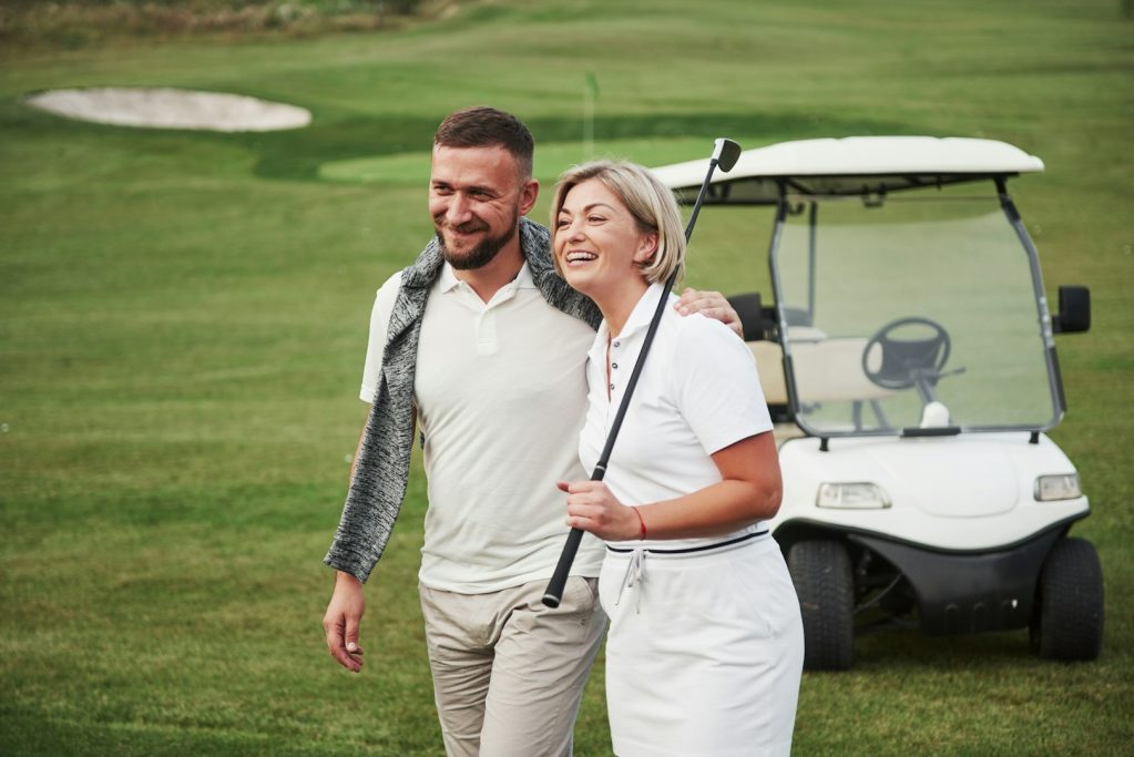 Pareja sonriente caminando por el campo de golf, con un carrito de golf al fondo.